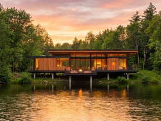 Maison contemporaine en bois sur pilotis surplombant un lac calme au coucher du soleil.